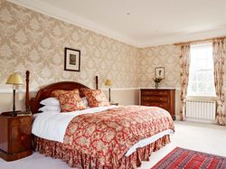 A luxurious bedroom with a wooden four-poster bed, rich red and gold bedding, patterned wallpaper, antique furniture, and a large window with floral curtains.