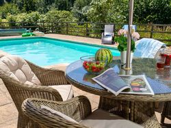 A relaxing poolside setting with wicker chairs, a glass table holding fresh fruit, drinks, and a magazine. A turquoise pool and lush greenery create a tranquil atmosphere.