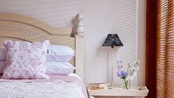 A cozy bedroom with a light wooden bed, pink floral bedding, and a matching cushion. A bedside table holds books, fresh flowers, and a lamp with a patterned shade.