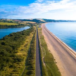 A stunning aerial view of a coastal road running between a calm blue lagoon and a golden sandy beach, with rolling green hills and a clear sky in the background.
