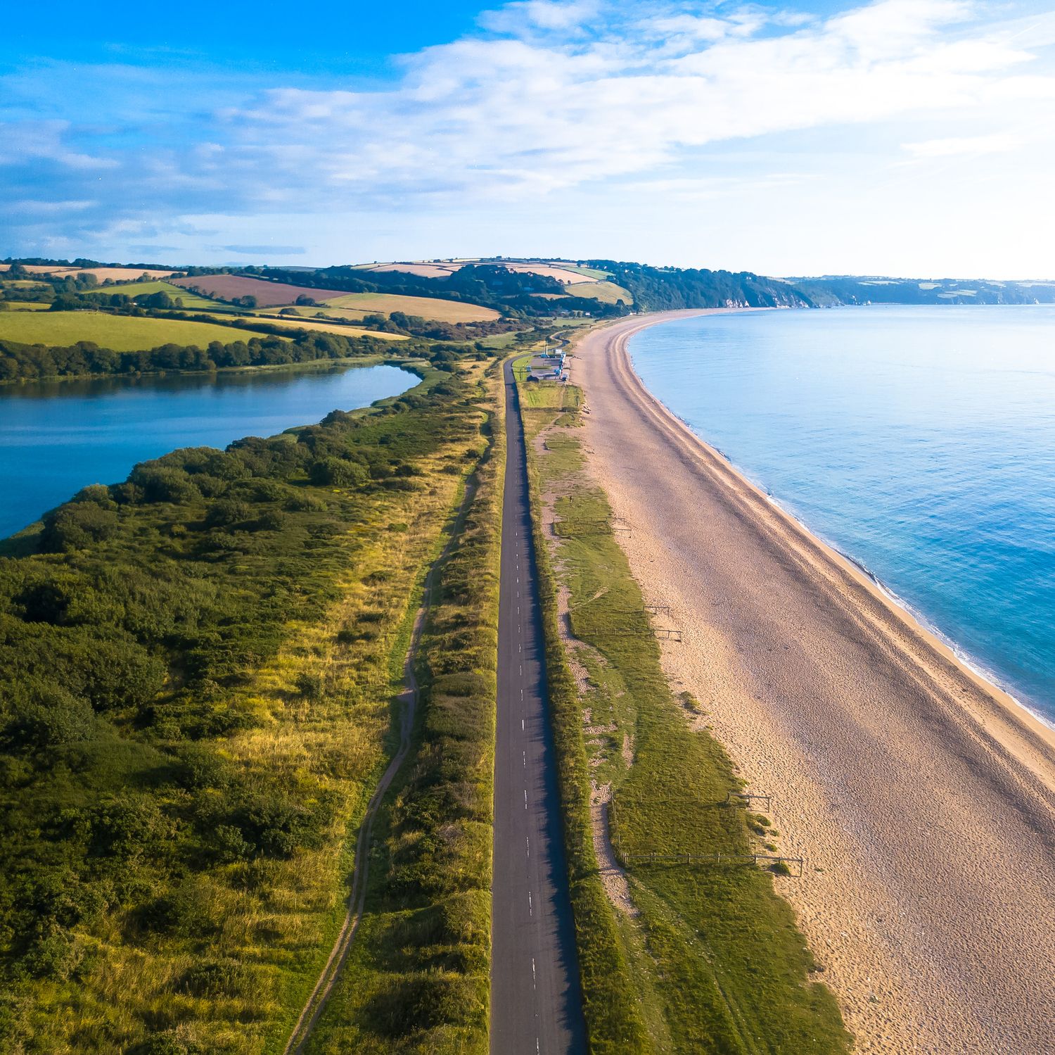 A stunning aerial view of a coastal road running between a calm blue lagoon and a golden sandy beach, with rolling green hills and a clear sky in the background.