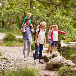 A family with three children explores a scenic forest trail by a river. The mother carries a backpack, the father has a child on his shoulders, and the kids look excited.
