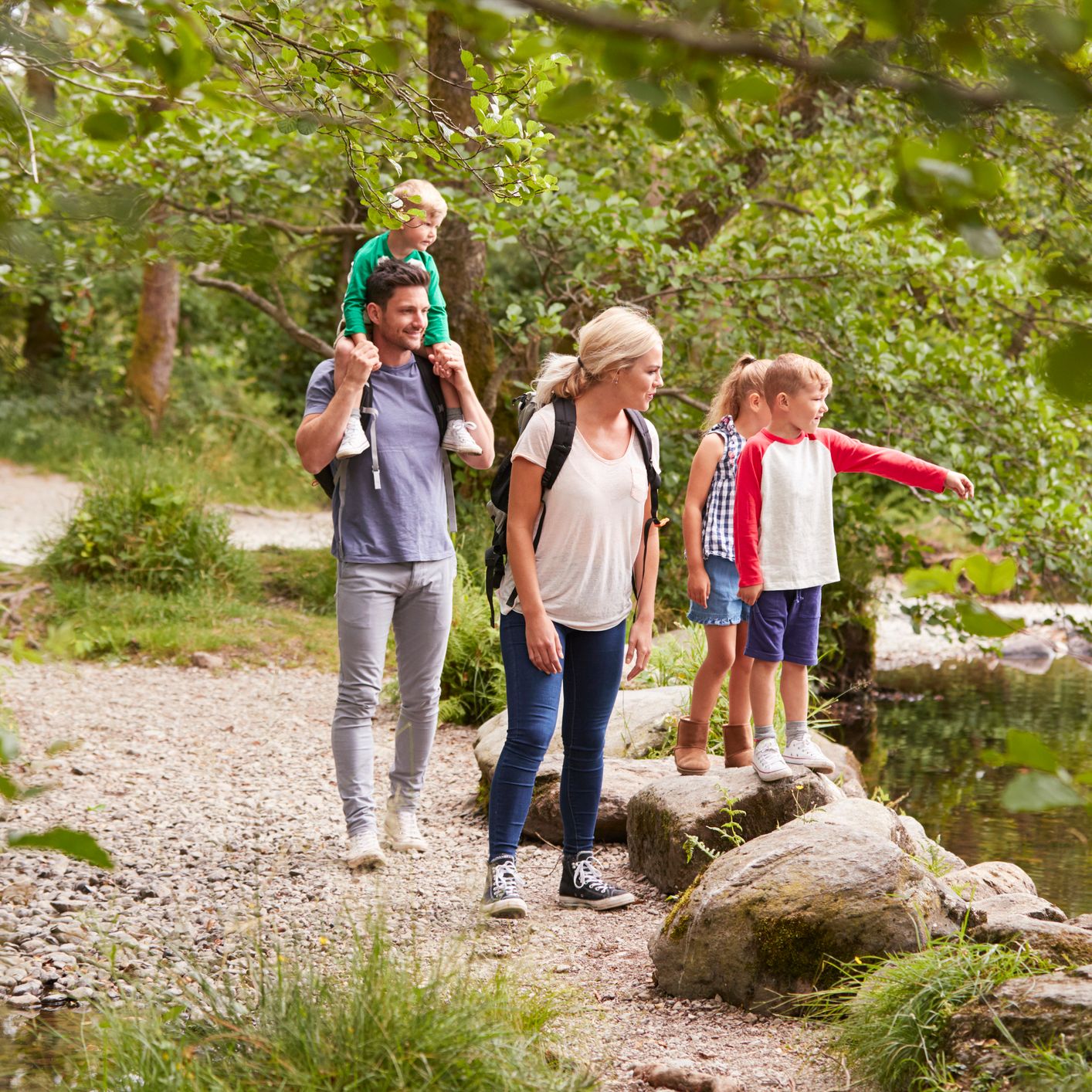 A family with three children explores a scenic forest trail by a river. The mother carries a backpack, the father has a child on his shoulders, and the kids look excited.