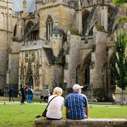 A couple sits on a stone ledge, admiring the architecture of a historic cathedral. People walk by, and pigeons scatter across the lush green lawn in the foreground.