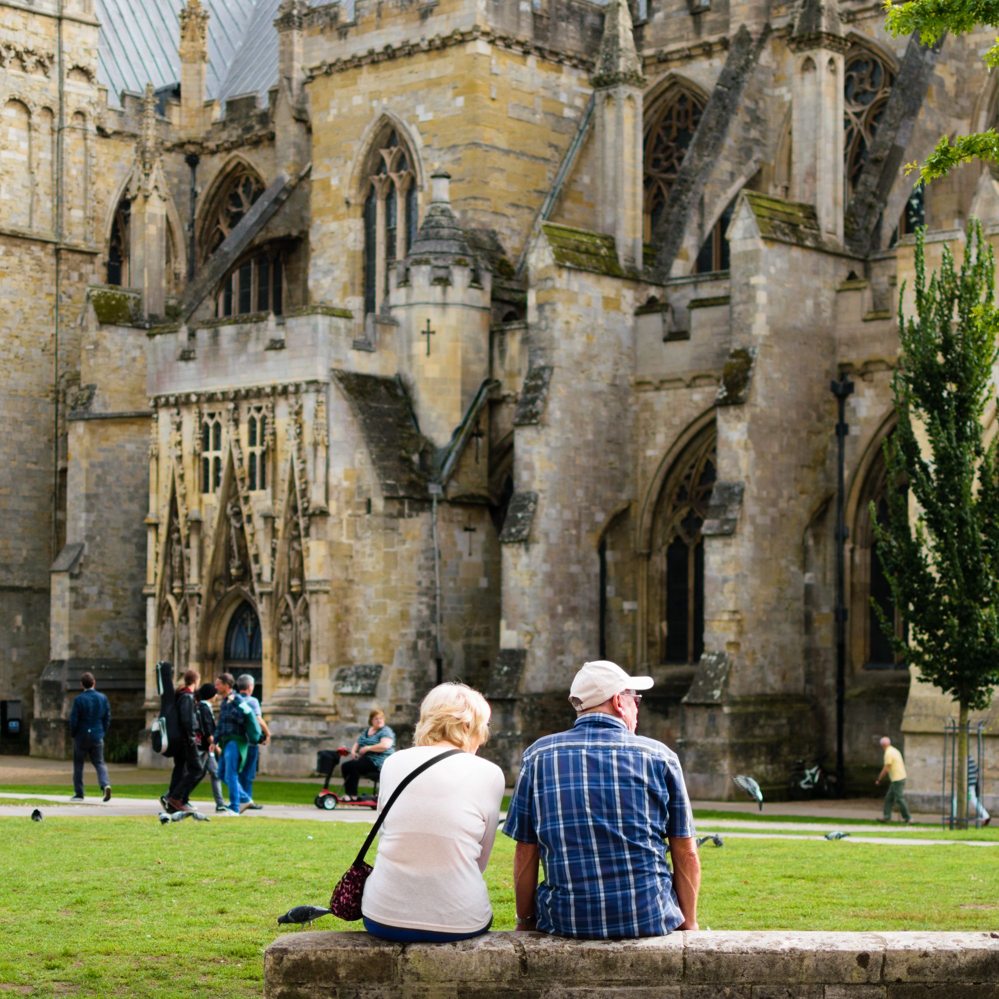 A couple sits on a stone ledge, admiring the architecture of a historic cathedral. People walk by, and pigeons scatter across the lush green lawn in the foreground.