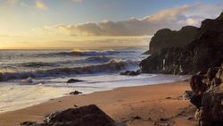 A scenic coastal view at sunset with waves crashing onto a sandy beach, surrounded by rugged cliffs and scattered rocks, bathed in golden light.