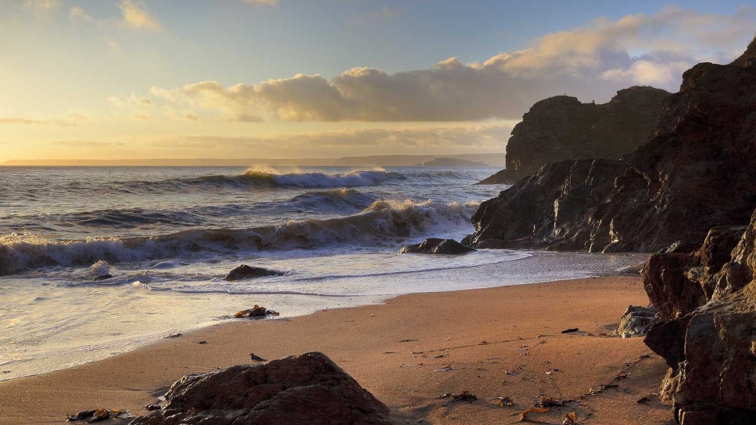 A scenic coastal view at sunset with waves crashing onto a sandy beach, surrounded by rugged cliffs and scattered rocks, bathed in golden light.