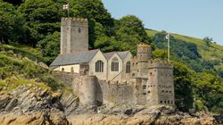 A historic stone castle with towers and arched windows sits on a rocky hillside by the water, surrounded by lush greenery, with flags flying under a clear blue sky.