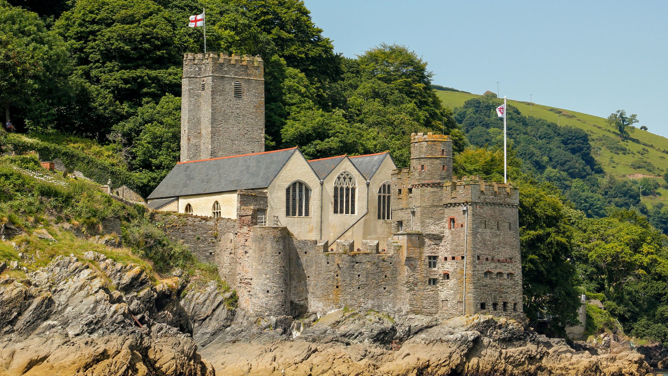 A historic stone castle with towers and arched windows sits on a rocky hillside by the water, surrounded by lush greenery, with flags flying under a clear blue sky.