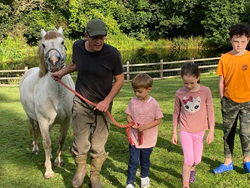A man in boots walks alongside a white pony, guiding a young boy holding the lead rope, while two other children walk beside them in a grassy field near a pond.
