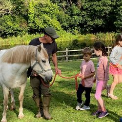 A man holds a red rope attached to a cream-colored horse while four young children stand nearby, watching attentively. Lush greenery and a pond are in the background.
