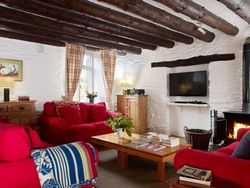 Cosy cottage living room with exposed wooden beams, red sofas, a wood-burning stove, rustic wooden furniture, and a TV mounted on a white stone wall.