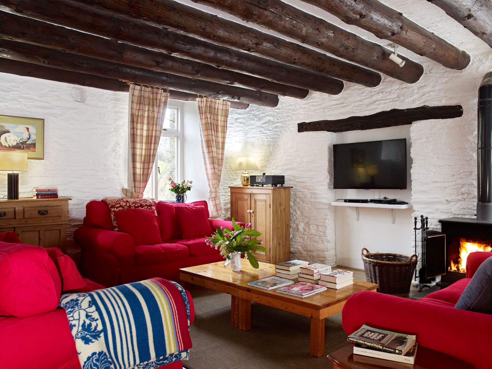 Cosy cottage living room with exposed wooden beams, red sofas, a wood-burning stove, rustic wooden furniture, and a TV mounted on a white stone wall.