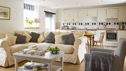 right open-plan cottage living space with a cream corner sofa, cosy cushions, a farmhouse-style kitchen, rustic wooden flooring, and a coffee table with books.