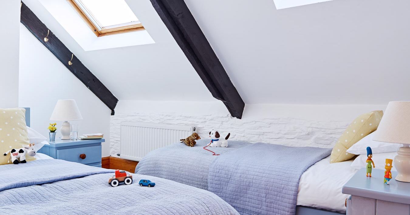 Cosy children's twin bedroom with sloped ceilings, skylights, soft lilac bedding, yellow polka dot cushions, wooden beams, and playful toys on the beds.