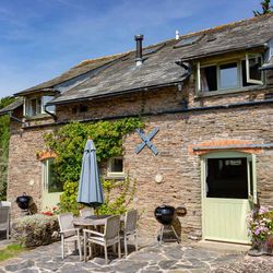 Charming stone cottage with ivy-covered walls, pastel green doors, outdoor dining tables with umbrellas, BBQ grills, and vibrant flower arrangements in a sunny courtyard.