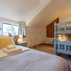Cosy cottage bedroom with a single bed and pastel blue bunk beds, soft quilted bedding, yellow polka dot cushions, rustic wooden accents, and a bright window view.