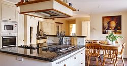 Traditional farmhouse kitchen with a large granite island, wooden dining table, built-in oven, rustic decor, and warm lighting for a cosy and inviting space.