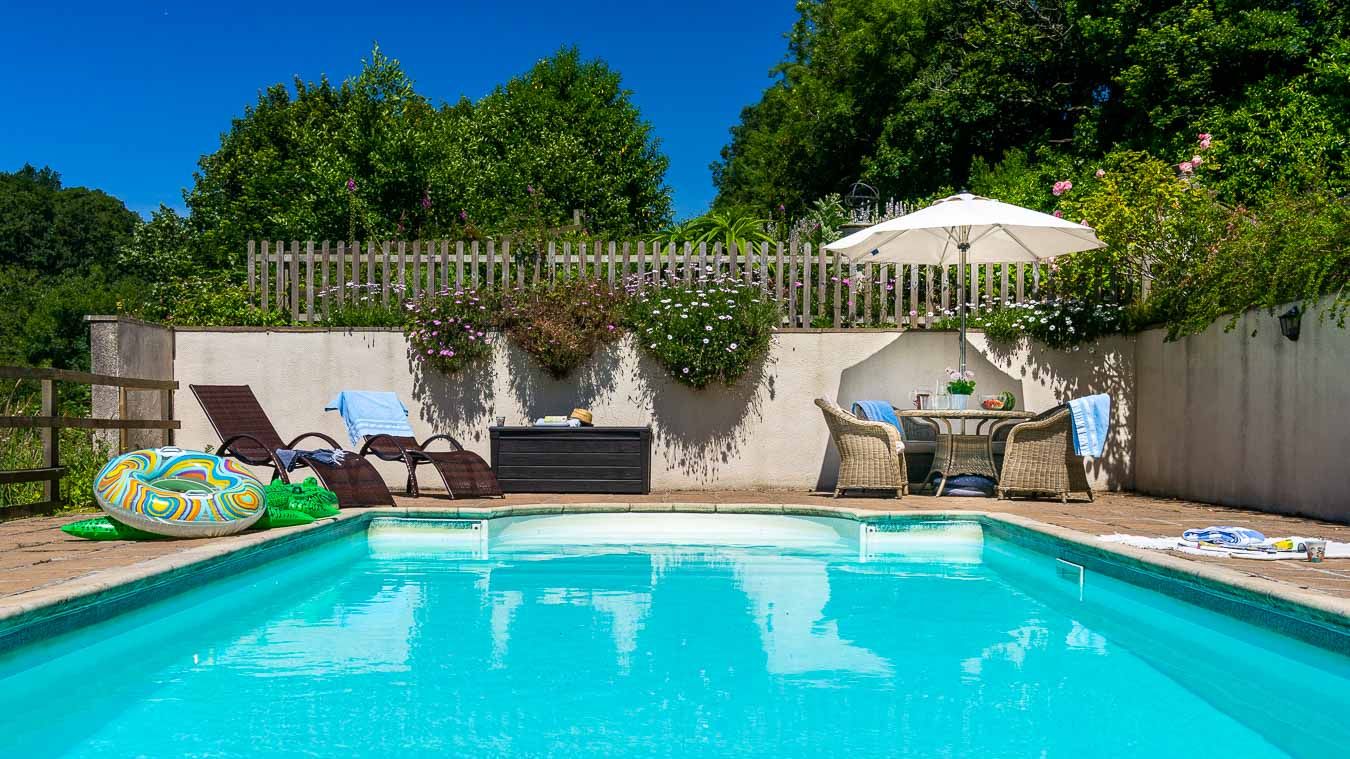 Outdoor swimming pool with clear blue water, sun loungers, parasol, and seating area surrounded by lush greenery and flowers on a sunny day.