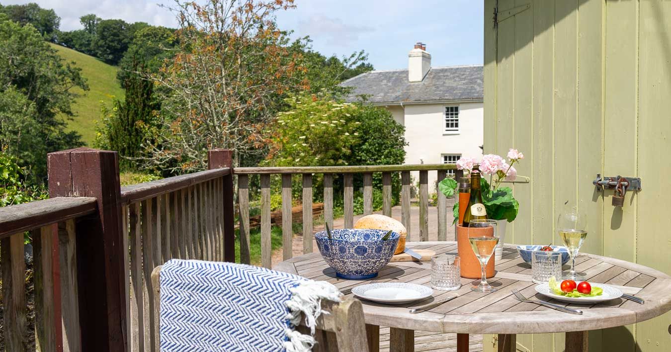 Scenic outdoor dining on a rustic wooden deck with a countryside view, featuring a round table set with wine, fresh bread, and salad beside a green garden shed.