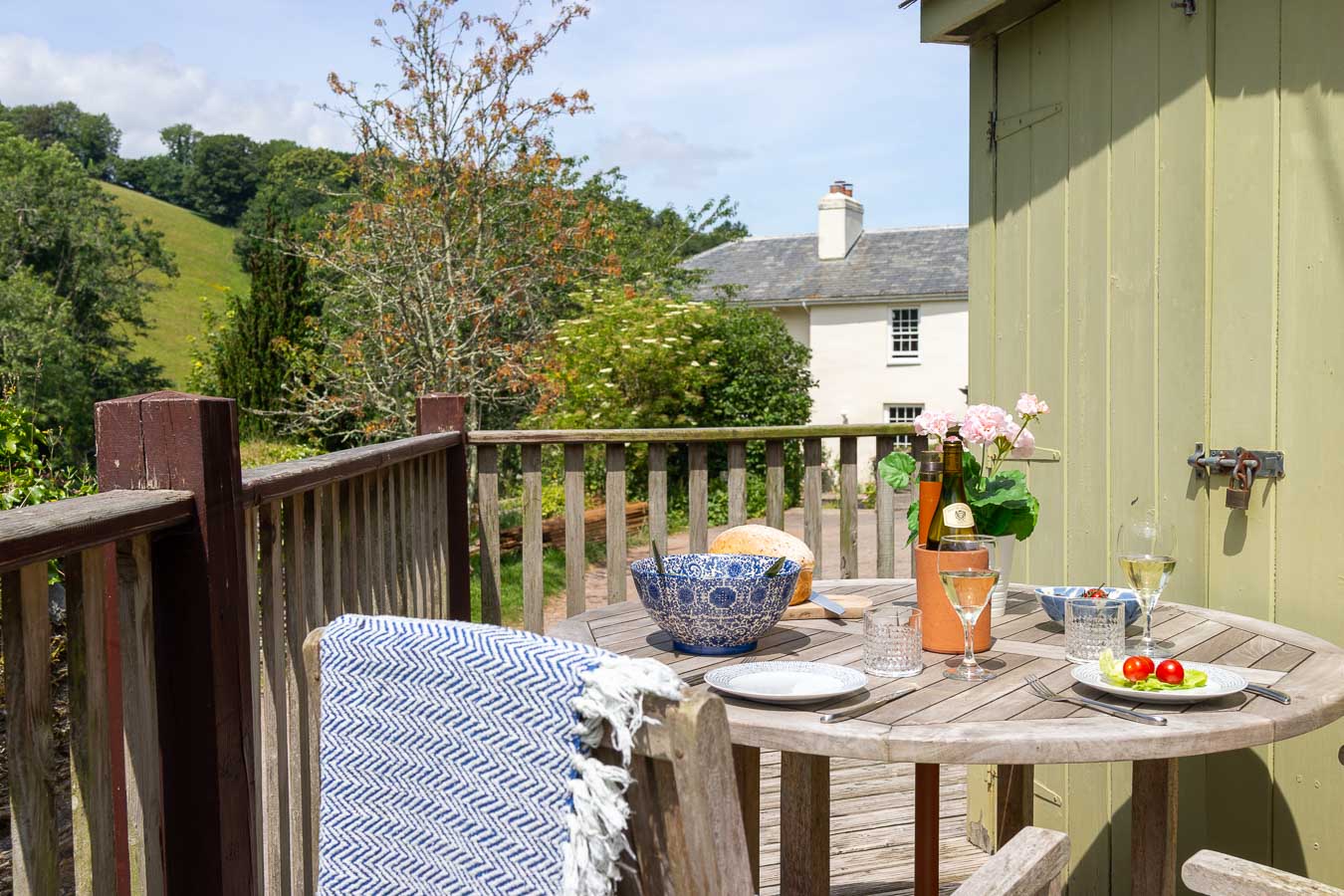 Scenic outdoor dining on a rustic wooden deck with a countryside view, featuring a round table set with wine, fresh bread, and salad beside a green garden shed.