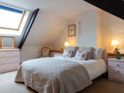 Charming attic bedroom with exposed beams, a cosy double bed with floral cushions, soft lighting, and a skylight allowing natural light to brighten the space.