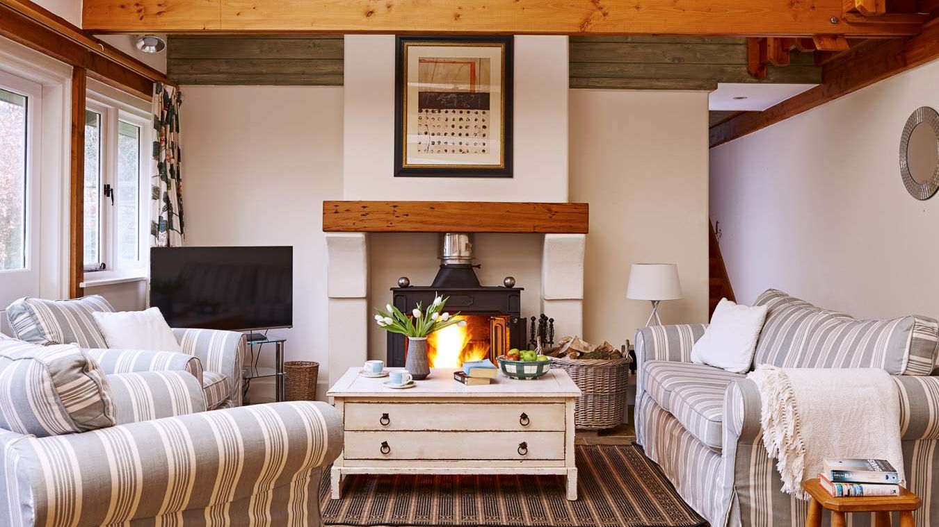 Cosy country-style living room with a wood-burning stove, striped sofas, wooden beams, and a rustic coffee table, creating a warm and inviting atmosphere.