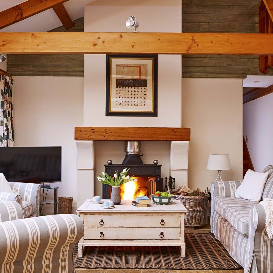 Cosy country-style living room with a wood-burning stove, striped sofas, wooden beams, and a rustic coffee table, creating a warm and inviting atmosphere.