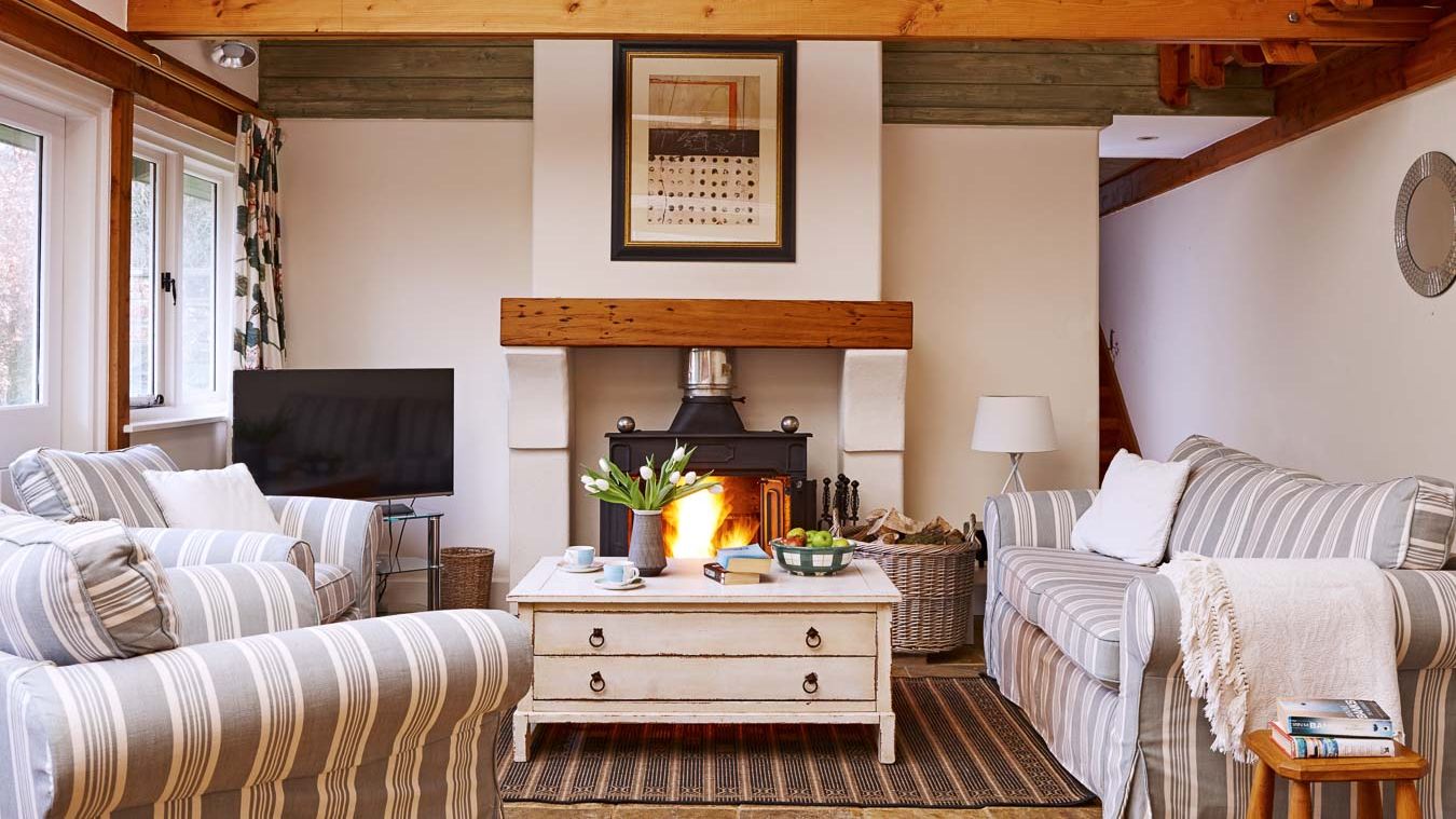 Cosy country-style living room with a wood-burning stove, striped sofas, wooden beams, and a rustic coffee table, creating a warm and inviting atmosphere.
