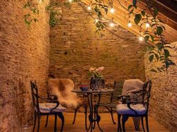 Cosy outdoor seating area under a rustic wooden lean-to with warm string lights, stone walls, and a bistro table set for drinks, creating a romantic evening ambience.