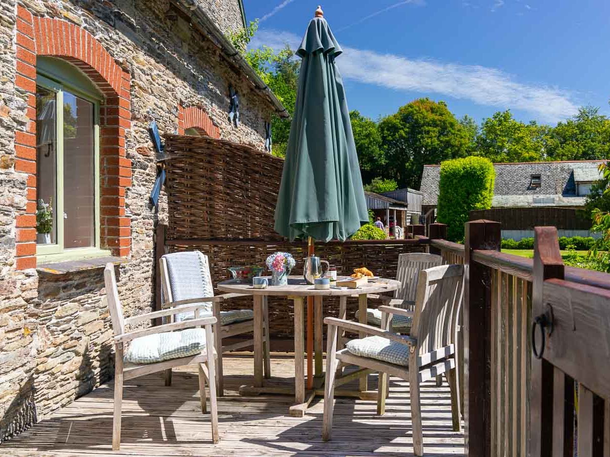 Charming outdoor deck with wooden furniture, a large green umbrella, and a rustic stone wall, set for a relaxing al fresco breakfast in the sunshine.