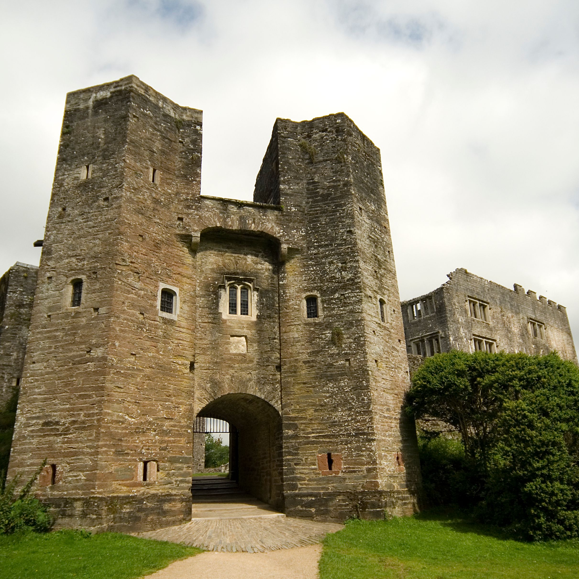 Ancient stone castle with a grand arched entrance, fortified towers, and partially ruined walls, surrounded by lush greenery under a cloudy sky.
