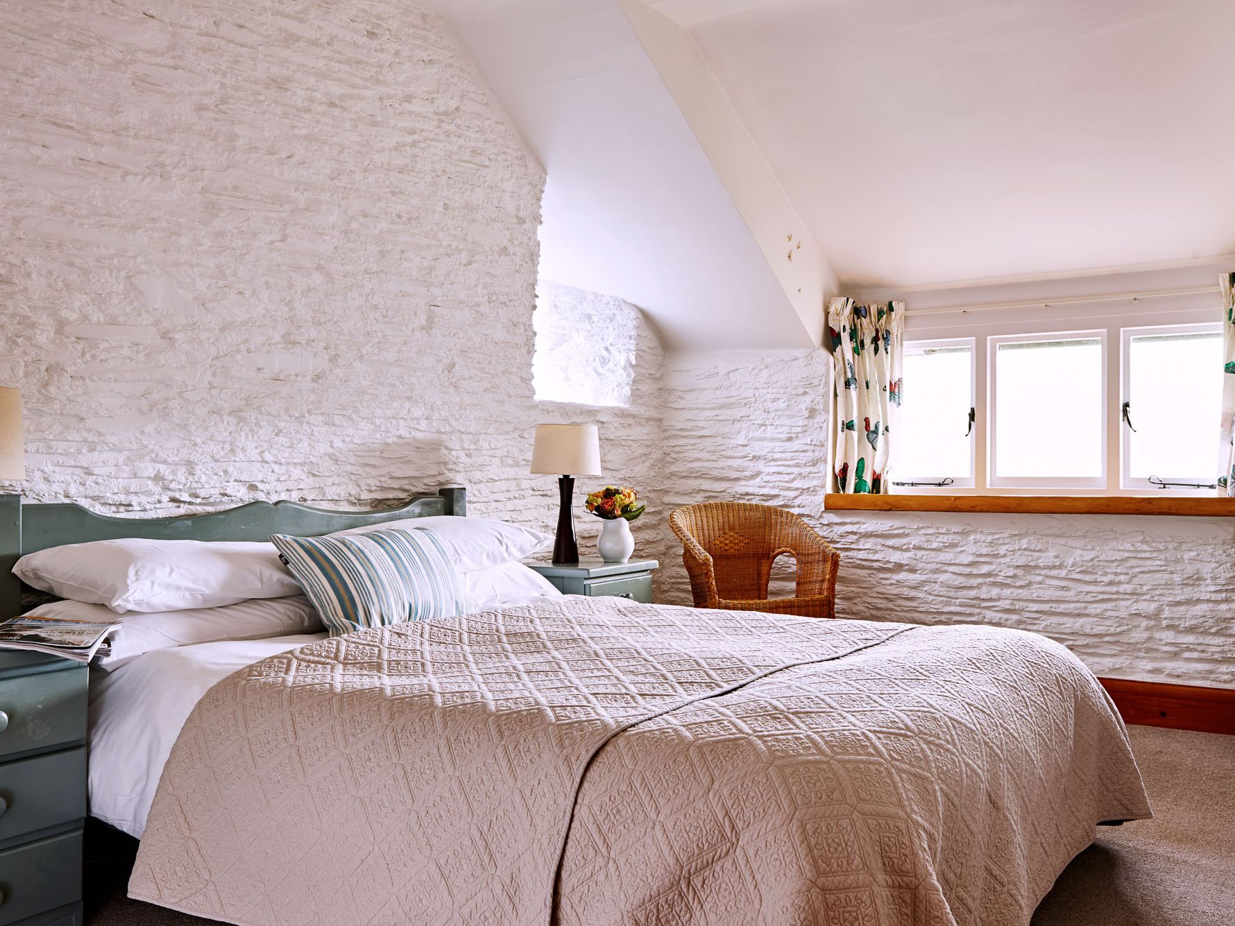 Cosy bedroom with a rustic green bed frame, white stone walls, soft beige bedding, a wicker chair, and a window with floral curtains letting in natural light.