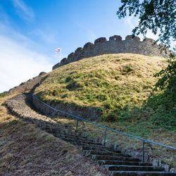 Stone steps winding up a grassy hill towards an ancient circular castle ruin with crenelated walls, a flag flying at the top, and a clear blue sky above.