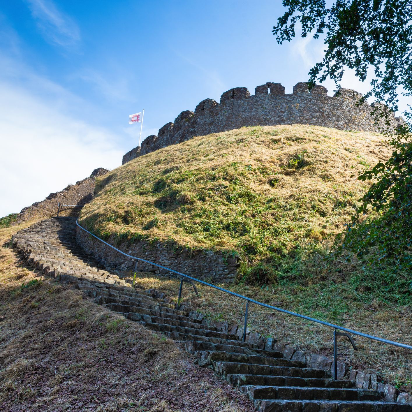 Stone steps winding up a grassy hill towards an ancient circular castle ruin with crenelated walls, a flag flying at the top, and a clear blue sky above.