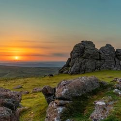 A scenic view of rugged granite rock formations on Dartmoor at sunset, with a golden sky, rolling hills in the background, and a grazing pony on the grass.