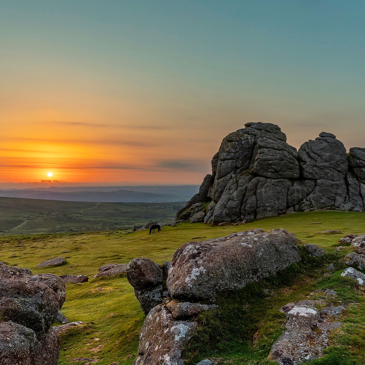 A scenic view of rugged granite rock formations on Dartmoor at sunset, with a golden sky, rolling hills in the background, and a grazing pony on the grass.