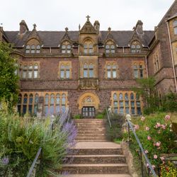 A historic Victorian-style building with ornate stonework, large windows, and a grand entrance, surrounded by lush gardens and colourful flowers.