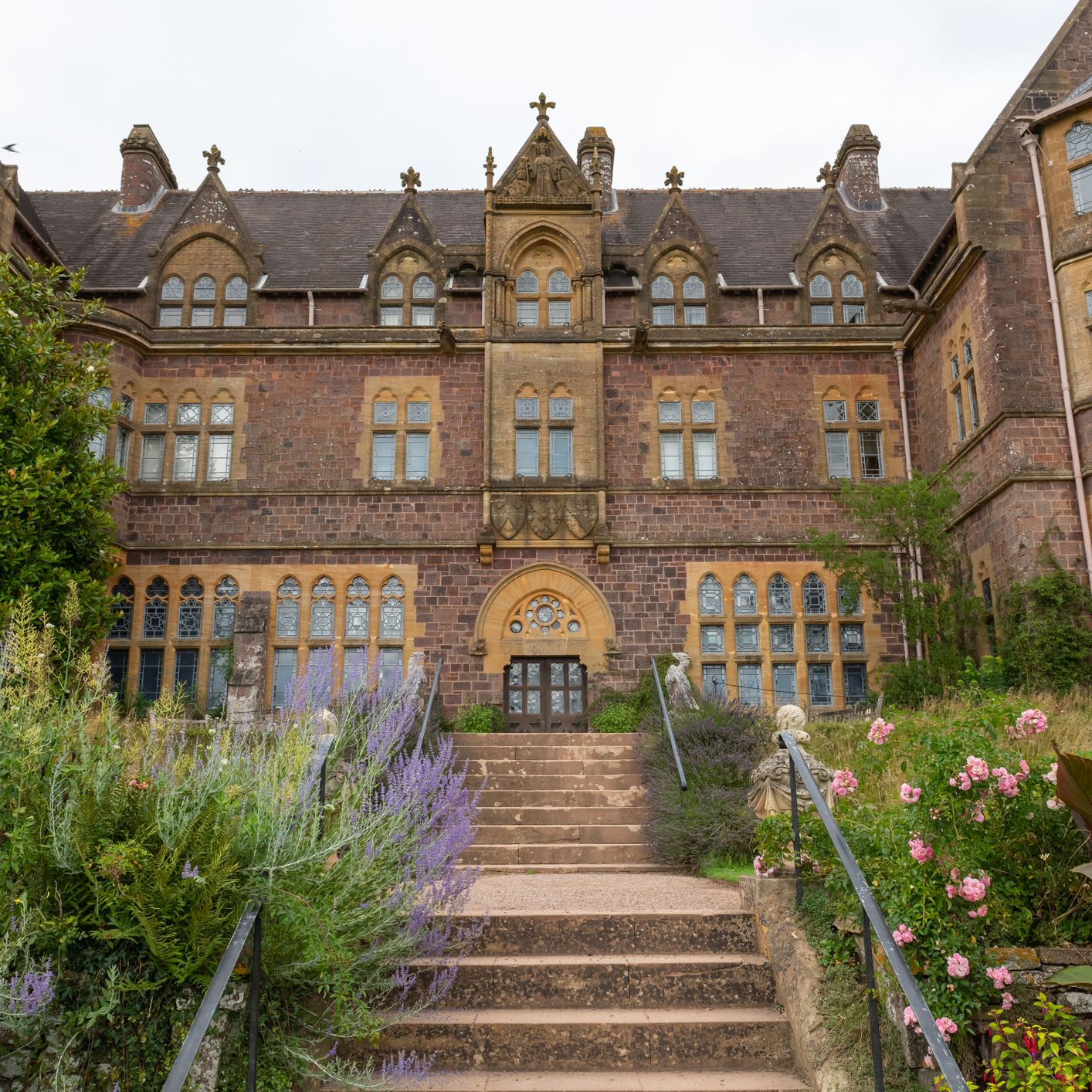 A historic Victorian-style building with ornate stonework, large windows, and a grand entrance, surrounded by lush gardens and colourful flowers.