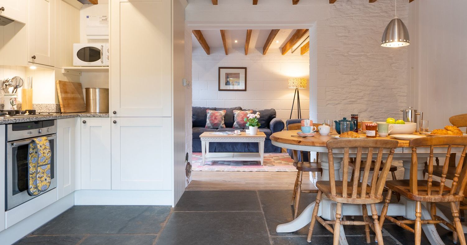 A cosy cottage kitchen and dining area with a wooden table set for breakfast, leading into a snug living space with a navy sofa, beamed ceiling, and soft lighting.