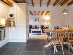 A cosy cottage kitchen and dining area with a wooden table set for breakfast, leading into a snug living space with a navy sofa, beamed ceiling, and soft lighting.