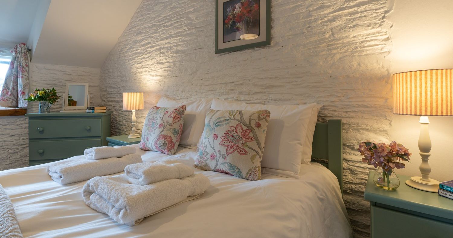 A charming cottage bedroom with a cosy double bed, floral cushions, soft lighting, and rustic white stone walls, creating a warm and inviting atmosphere.