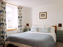 A bright and airy cottage bedroom with a cosy double bed, striped cushions, soft lighting, and rustic white stone walls, complemented by butterfly-patterned curtains.