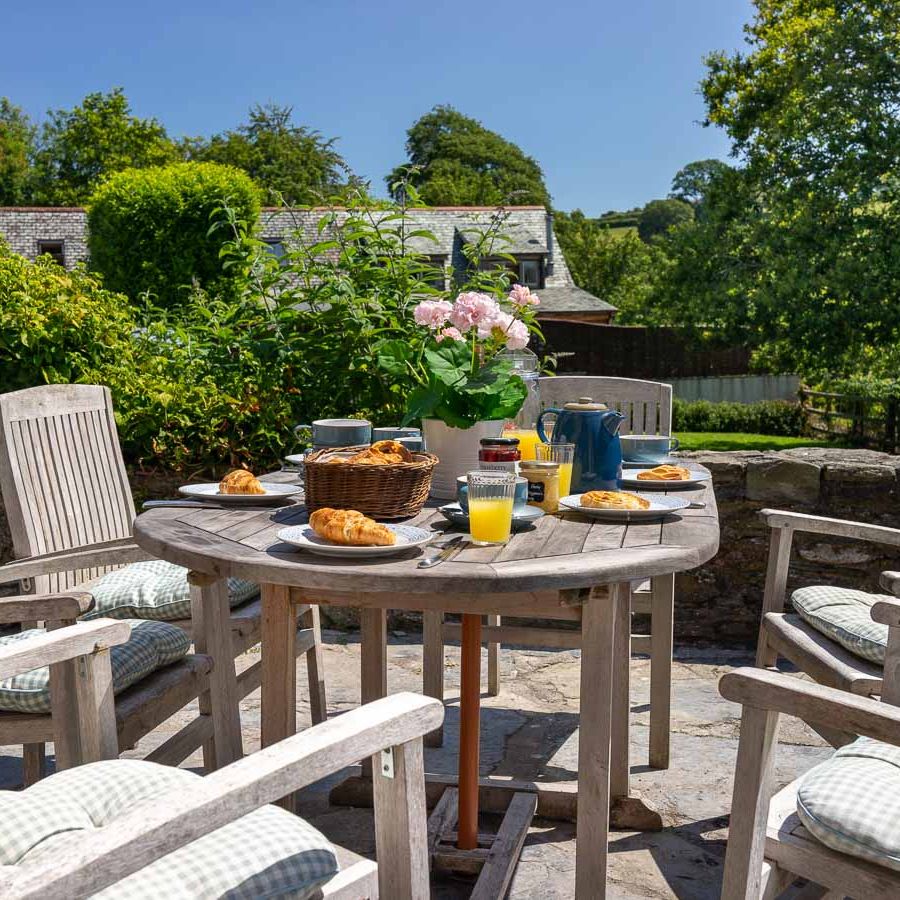 A scenic outdoor breakfast setup with wooden chairs and a round table, featuring croissants, juice, and coffee, surrounded by lush greenery and a countryside view.