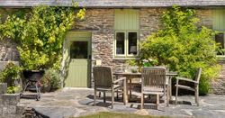 A charming stone cottage with green doors and windows, an outdoor dining area, and a barbecue, surrounded by lush greenery and bathed in sunlight.