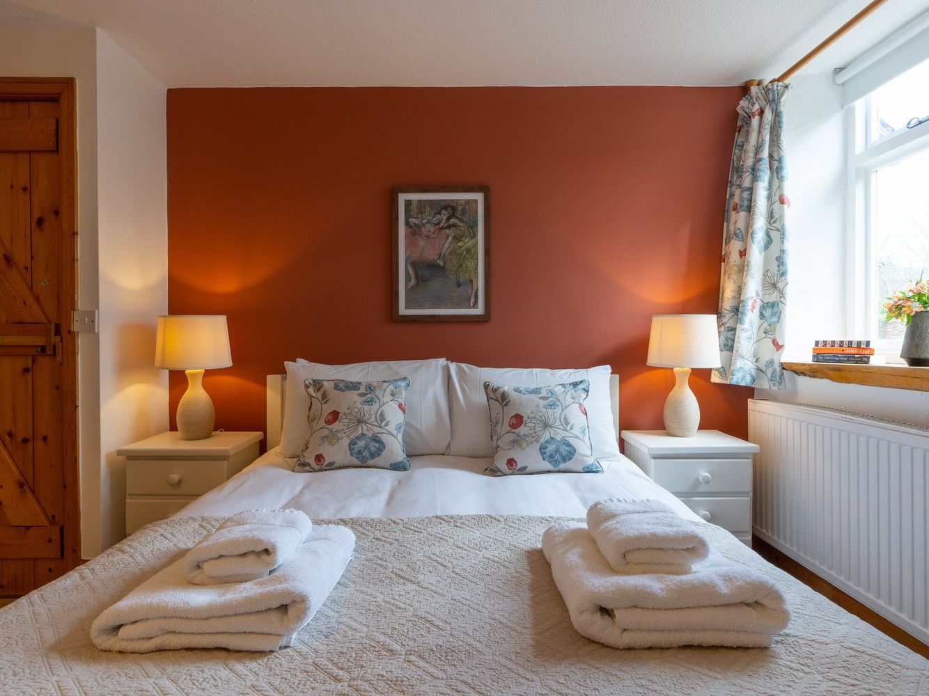 A cosy cottage bedroom with a double bed, warm lighting, a rustic wooden door, and a terracotta accent wall, creating a welcoming and relaxing atmosphere.
