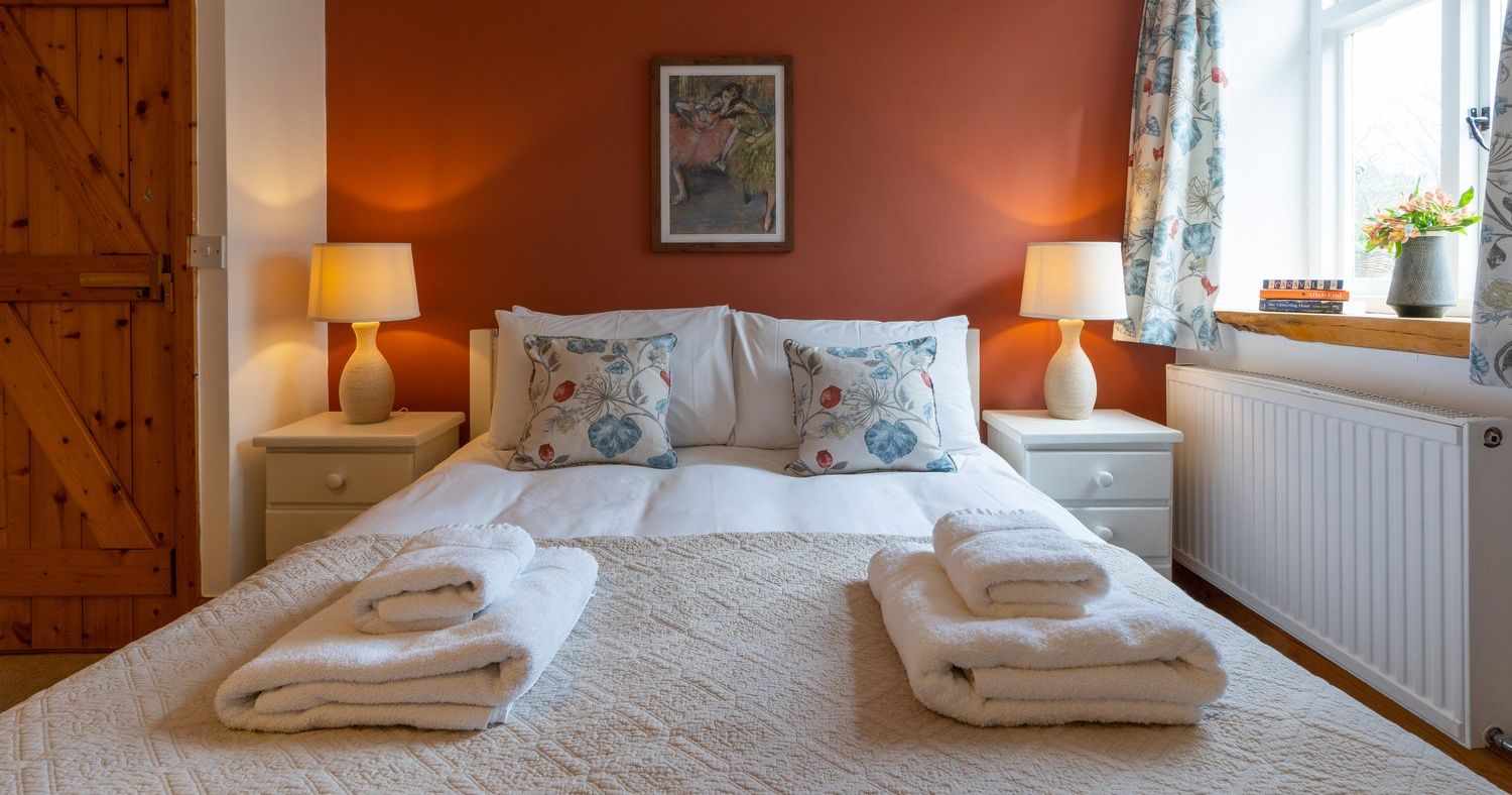 A cosy cottage bedroom with a double bed, warm lighting, a rustic wooden door, and a terracotta accent wall, creating a welcoming and relaxing atmosphere.
