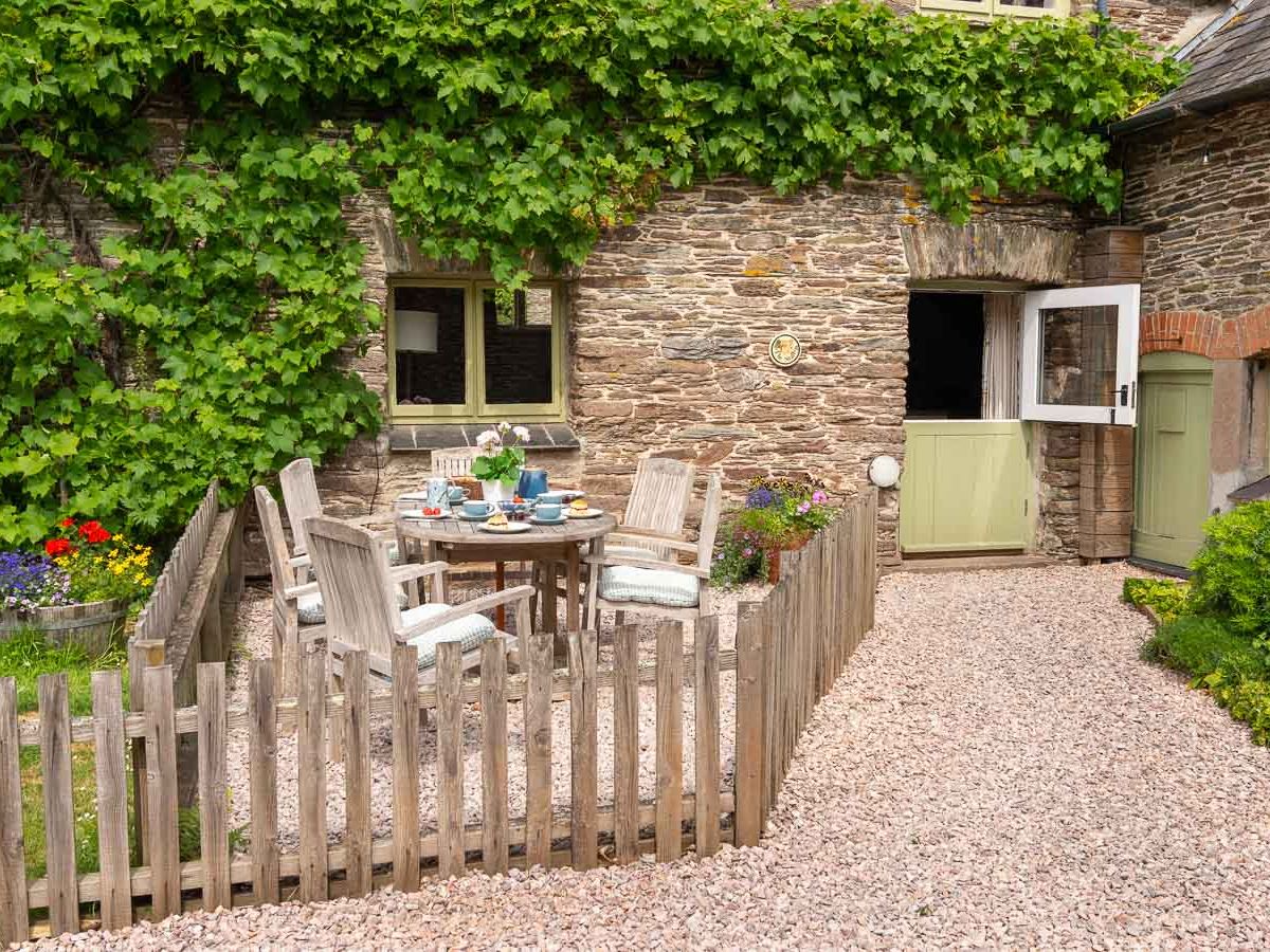 A charming stone cottage with a vine-covered wall, a quaint enclosed garden, and an outdoor dining area, featuring rustic wooden furniture and colourful flowers.