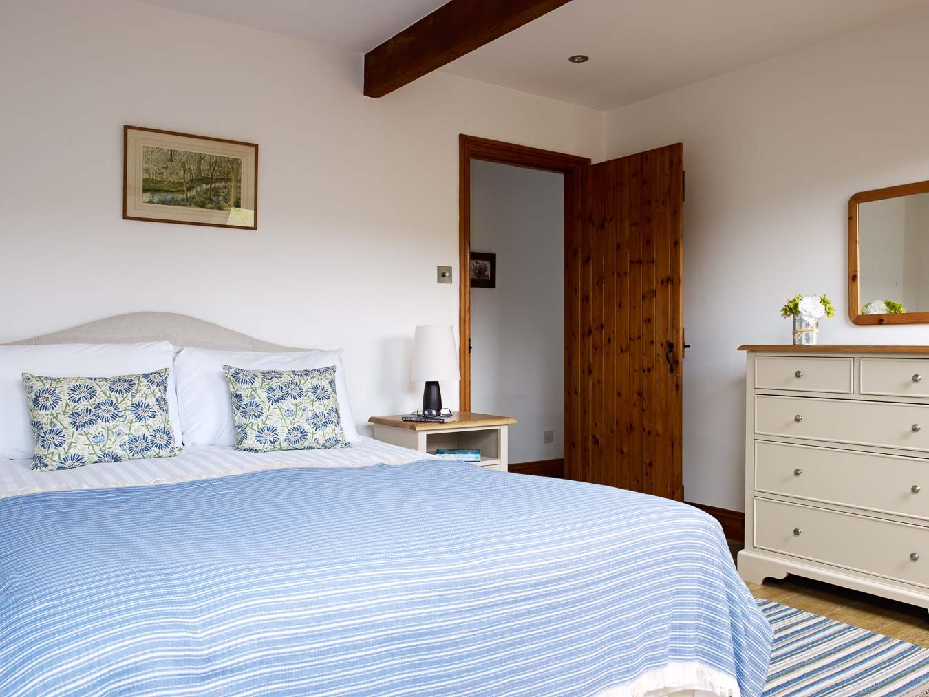Bright and airy bedroom with a king-size bed, blue and white striped bedding, floral cushions, wooden furniture, and a cosy, rustic charm.