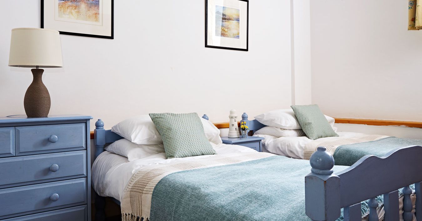 Charming twin bedroom with pastel blue wooden beds, soft green cushions, matching throws, and nautical-themed artwork, creating a cosy and relaxing atmosphere.